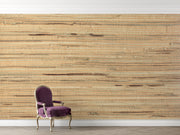A large-scale woven wall mural with a natural jute or seagrass texture in warm beige and brown tones, shown in a room setting behind a purple velvet armchair.