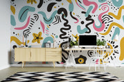A colorful abstract wavy patterned wallpaper displayed on a wall in a modern home office setting, featuring a light wood sideboard, a white desk with a computer, and a black and white chevron rug.