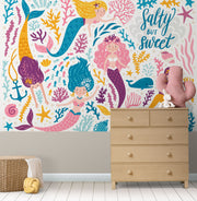 Colorful mermaid-themed wall mural in a children's playroom, featuring smiling cartoon mermaids with vibrant tails, sea creatures and coral.