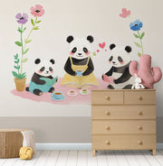 A children's room featuring a playful wall mural of a panda family having a tea party, with pastel-colored flowers and cozy decor including a wooden dresser and plush toys.