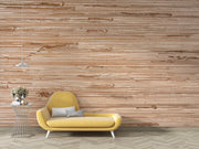 A modern yellow chaise lounge chair with two gray pillows, positioned next to a wire side table with a vase, set against a large textured woven wall panel and light wood herringbone flooring.