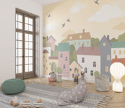Whimsical village wall mural featuring pastel houses, trees, clouds, and flying birds, styled in a cozy and creative kids’ playroom.