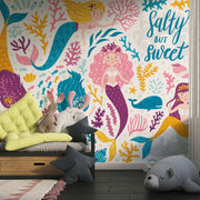 Colorful mermaid-themed wall mural in a children's playroom, featuring smiling cartoon mermaids with vibrant tails, sea creatures and coral.