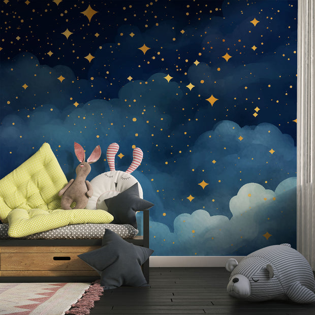 Starry night wall mural with golden stars and deep blue watercolor-style clouds, decorating a cozy bedroom with patterned bedding, soft toys and storage.